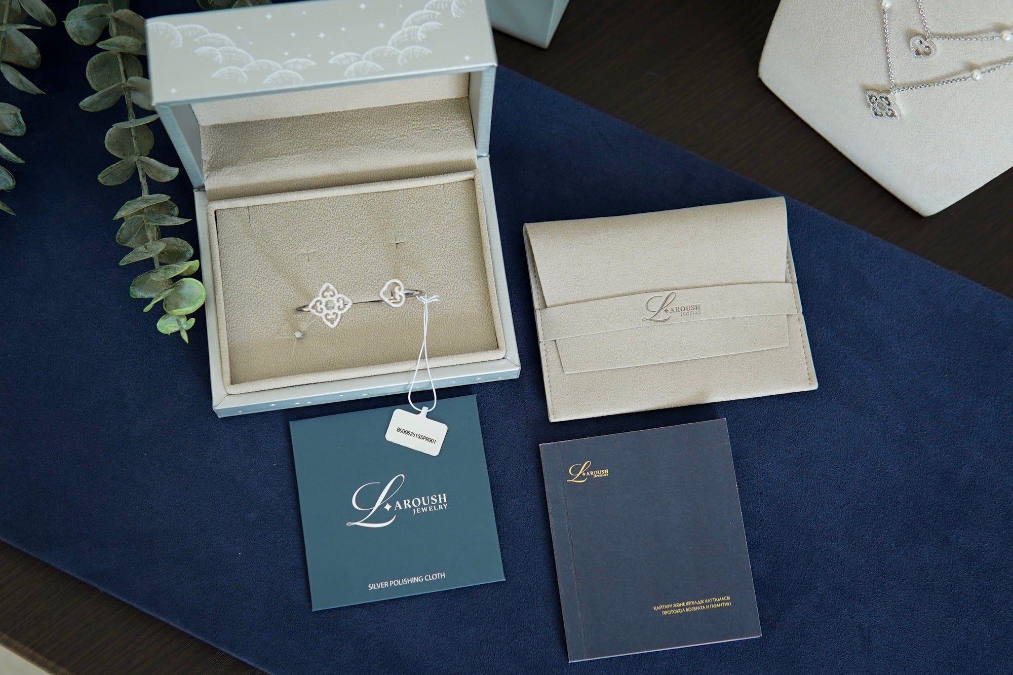 A jewelry set from Lamouch Paris, featuring a silver geometric charm bracelet and matching necklace displayed in an open light blue jewelry box, alongside a cream envelope, a dark blue brand card, and a dark blue booklet on a dark blue velvet surface.