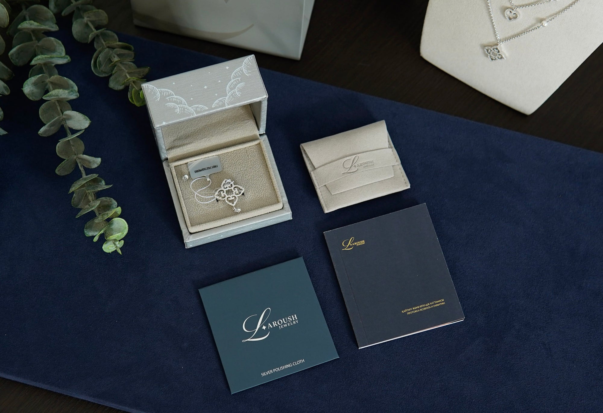 A jewelry gift set from L'Amour Paris, featuring a silver-toned necklace with a floral pendant in an open white box, a cream card case, a navy blue booklet, and a dark slate blue card, all arranged on a dark blue velvet surface with a eucalyptus sprig.